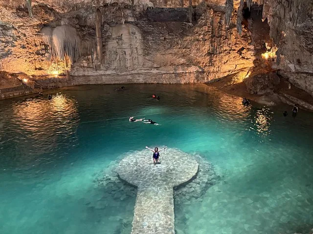 A person stands on the platform in Cenote Suytun with others swimming nearby.
