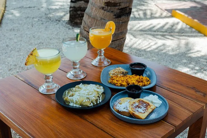 A wooden table with Mexican dishes, cocktails, and drinks outdoors.
